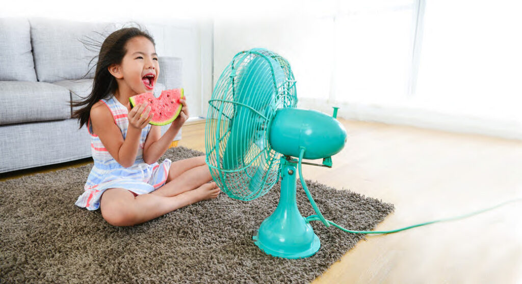 young girl holding watermelon sitting on tan shag rug on floor in front of a teal green electric fan.