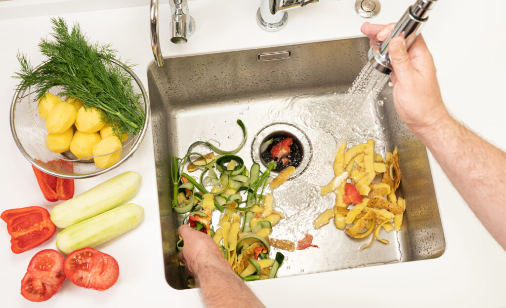 A person puts vegetable peels down the garbage disposal in a stainless steel kitchen sink, while running the faucet.